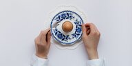 Zwei Hände neben einem gekochten Ei in einem weißblauen Ei-Service