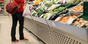 Eine Person, von der nur die untere Hälfte zu sehen ist, steht vor einem Gemüseregal im Supermarkt. In der Hand hat die Person eine Plastiktüte mit Äpflen darin
