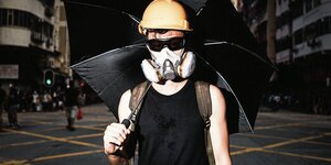 Eine Person trägt Atemschutzmaske, Sonnenbrille, einen gelben Helm und einen schwarzen Regenschirm
