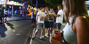 Im Vordergrund hält eine Frau einen mit Bier gefüllten Plastikbecher, im Hintergrund diskutieren junge Männer.