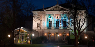 Das Deutsche Theater in Göttingen bei Nacht. Es es ist ein Haus mit spitzem Dach in der MItte. Links vom Gebäude aus Stein steht ein Anbau aus Glas, der hell erleuchtet ist. Das gesamte Gebäude wird von außen bunt angestrahlt.