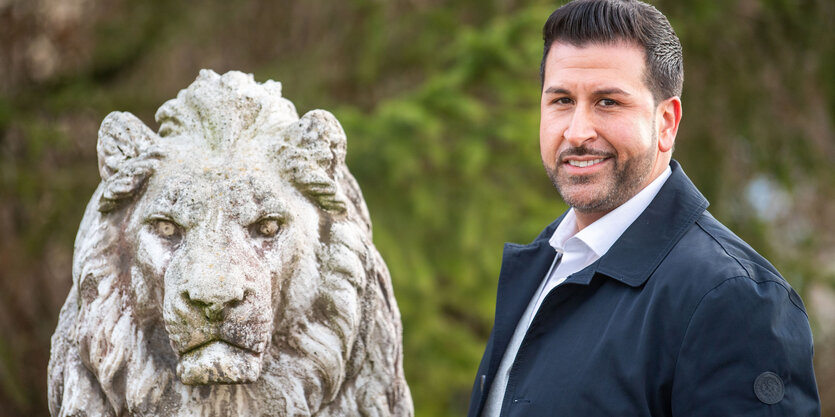 Ozan Iyibas steht rechts. links neben ihm die Statue eines Bayerischen Löwens. Beide blicken in die Kamera. Ozan Iyibas ist ein Mann mittleren Alters mit kurzen dunklen Haaren.