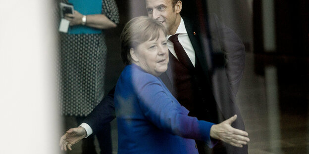 Angela Merkel und Emmanuel Macron strecken die Hand aus, aber in verschiedene Richtungen