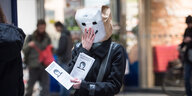 Person mit Papiertüte auf dem Kopf, auf die ein Gesicht gemalt ist, verteilt Flyer in einer Bahnhofshalle