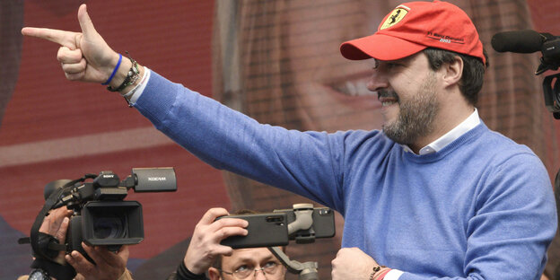 Salvini in Wollpulli und Ferrari-Cap hebt einen Arm, im Hintergrund Journalisten