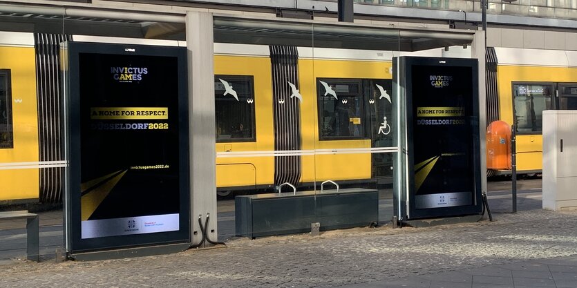 Eine Sitzbank aus Metall an einer Haltestelle, zwischen den Plätzen sind Metallbügel. Dahinter parkt eine gelbe Tram