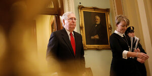 Mitch McConnell geht durch den Senat, neben ihm ein Porträt und eine Frau, die Akten trägt.