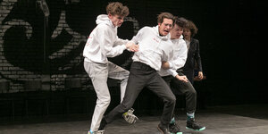 Vier junge Männer auf einer Bühne, alle in Bewegung, der in der Mitte wird bedrängt.