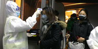 Reisende mit Atemschutzmasken werden von einer Person im Schutzanzug auf erhöhte Temperatur untersucht