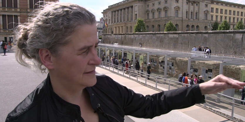 Eine Frau steht im Freien und zeigt auf tiefer liegende Mauerrest.