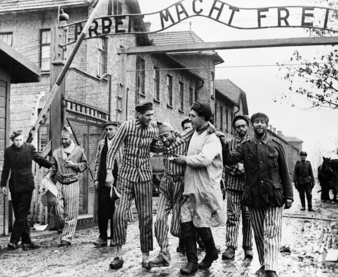 Insassen des Konzentrationslagers Auschwitz-Birkenau in schwarz-weißer Häftlingskleidung laufen bei der Befreiung unter dem Tor mit der Aufschrift "Arbeit macht frei" hindurch