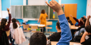 Ein Schüler meldet sich, die Lehrerin steht mit dem Rücken zu ihm an der Tafel