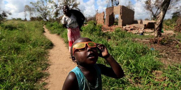 Ein Junge mit Sonnenbrille steht vor einer zerstörten Hütte, die durch den Zyklon Idai zerstört wurde.
