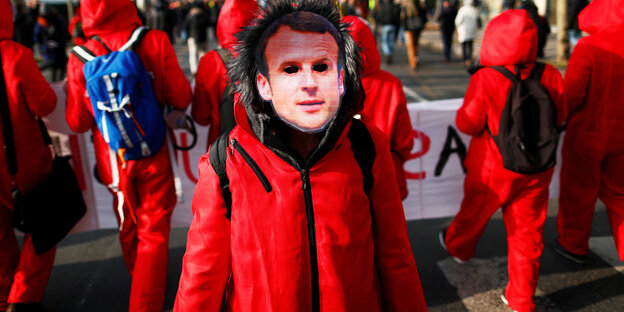 Demonstranten in Paris tragen rote Overalls. Auf einem klebt das Gesicht des französischen Präsidenten Emmanuel Macron