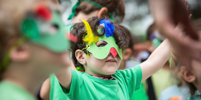 Ein Junge trägt eine bunte Maske auf dem Gesicht und streckt beide Arme in die Luft