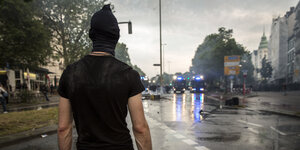 Ein Mann im schwarzen T-Shirt und Hasskappe schaut eine regennasse Straße hinunter, am Horizont Wasserwerfer