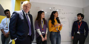 Boris Johnson steht grübelnd vor einer Tafel mit Rechenaufgaben, hinter ihm Studenten