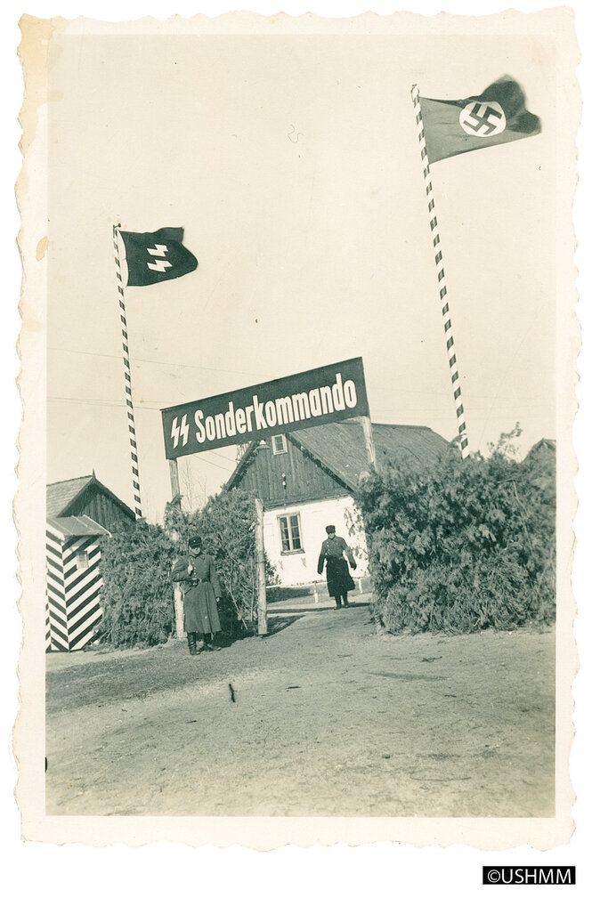 Sonderkommando steht es mit weißen Buchstaben auf einem schwarzem Grund. Zu beiden Seiten weht eine schwarze Fahnen mit SS-Zeichen und Hakenkreuz. Der Zaun ist mit Kiefernzweigen verblendet