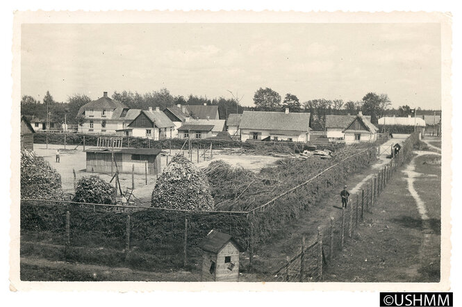 Neben der Baracke am linken Bildrand und im Vordergrund zwischen den Brennholz-Stapeln ist jeweils ein jüdischer Zwangsarbeiter zu erkennen.