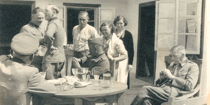 Acht Personen, sechs Männer und zwei Frauen, haben es sich im Freien auf einer Terrasse gemütlich gemacht, auf dem Tisch an dem sie sitzen stehen teils hochwertige Kristallgläser, wahrscheinlich aus dem Besitz der Ermordeten