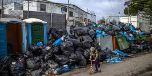 Ein kleines Kind fährt auf einem Tretroller, im Hintergrund sind Dixi-Toiletten und Müllberge zu sehen