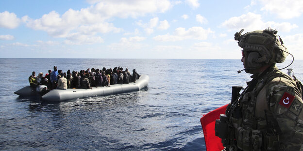 Migranten auf einem Schlauchboot im Mittelmmer, im Vordergrund auf einem Kriegsschiff ein Türkischer Soldat.