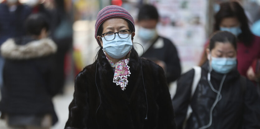 Eine Passantin in Hongkong mit Atemmaske. Im Hintergrund unscharf weitere PassantInnen mit Atemmasken.