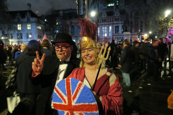 eine Frau mit römischer Kopfbedeckung und einem Schutzschild mit Union Jack, daneben ein Mann mit schwarzem Anzug und Melone. Sie stehen auf dem Parliament Square, spätabends, im Hintergrund einer Menschenmenge