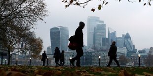 Menschen spazieren in einem Park vor den Hochhäusern des FInanzviertels in London.