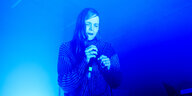 In blaues Licht getaucht, die Augen geschlossen, sieht man eine Frau mit langen Haaren und Mikrofon.
