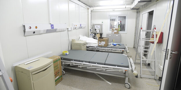 Im provisorisch eingerichteten Krankenzimmer stehen zwei Betten und eine Leiter. Die Wände sind kahl und das Licht ist künstlich und grell.