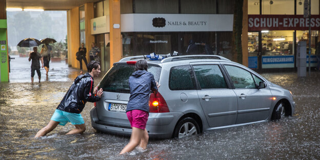 Zwei Personen schieben ein Auto bei Hochwasser und strömenden Regen