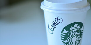 Hand greift nach Starbucks-Becher, darauf steht "James" mit Marker geschrieben (Screenshot)