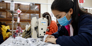 Seitenasicht einer Arbeiterin an der Nähmaschine in einer Textilfabrik trägt eine Mundschutzmaske