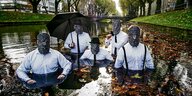 Fünf Männer mit Masken und weißen Hemden waten durch einen Kanal.