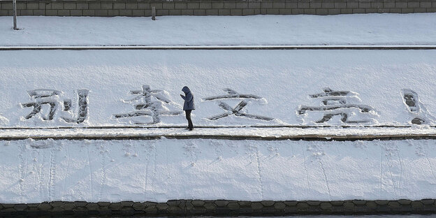 Großflächige chinesische Schriftzeichen, die in den Schnee geschrieben worden sind. In der Mitte steht ein Mann