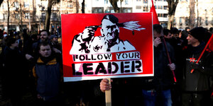 auf einer Demo hält ein Mensch ein Schild hoch, auf dem sich Adolf Hitler in die Schläfe schießt, darunter steht: „Follow Your Leader“
