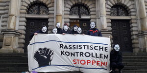 Menschen mit Pappmasken und -nasen stehen vor dem Hamburger Justizgebäude. Sie halten ein Plakat auf dem steht: "Rassistische Kontrollen stoppen"