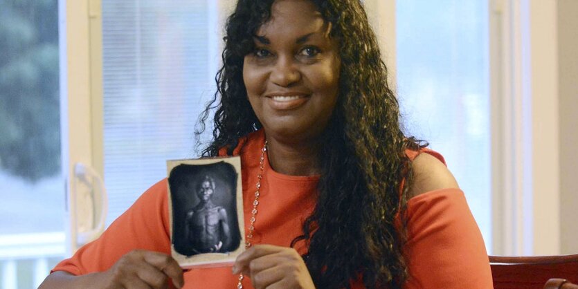 Eine junge Frau sitzt an einem Tisch und hält ein kleines schwarzweißes Foto hoch, auf dem ein Mann unbekleidet zu erkennen ist