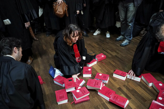 Rote Bücher liegen auf dem Parkettboden, Anwälte in schwarzen Roben stehen drum herum, eine kniet neben den Büchern