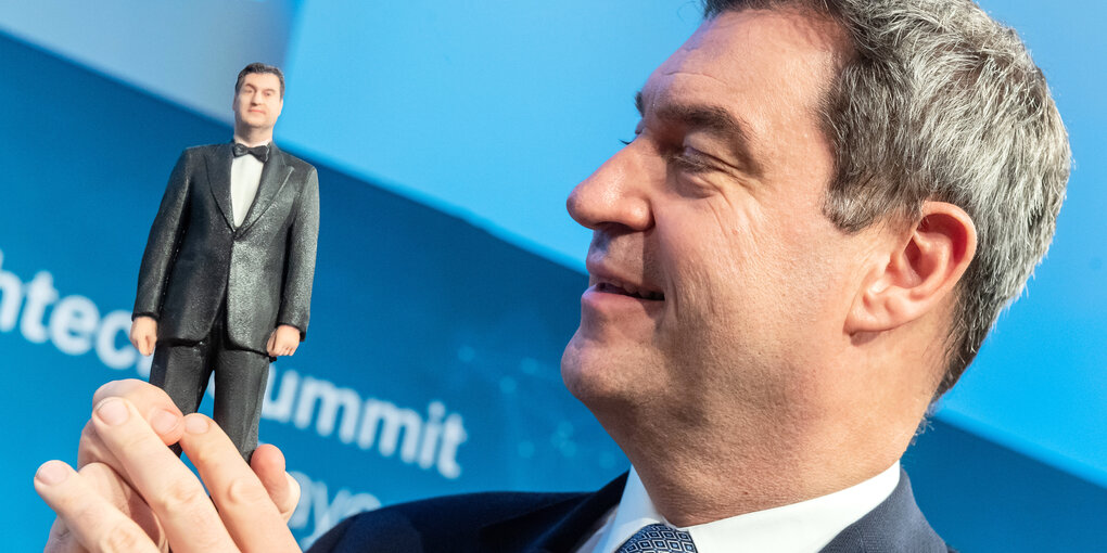 Markus Söder schaut eine figur vonb sich selber an.