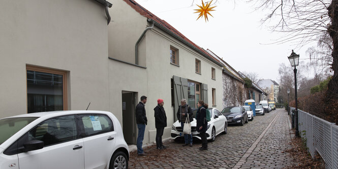 Vier Menschen stehen am Straßenrand neben einem niedrigen weißen Haus und unterhalten sich