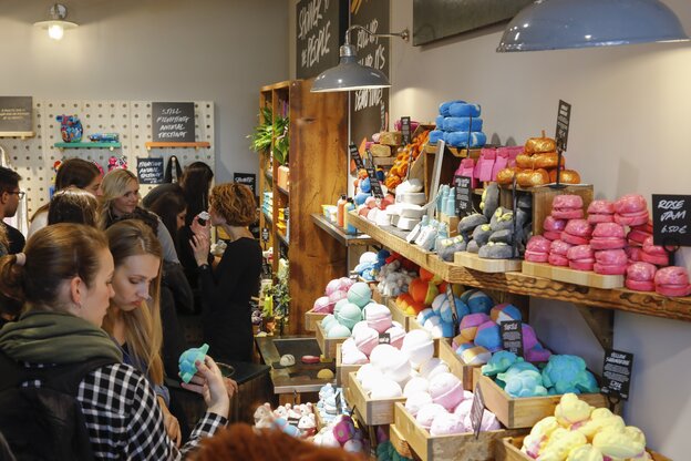 junge Menschen drängen sich vor dicken Holzregalen mit Bergen von unverpackten, bunten Badekosmetika junge Menschen drängen sich vor dicken Holzregalen mit Bergen von unverpackten, bunten Badekosmetika