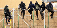 Migranten klettern über einen Zaun und werden von einem Spanischen Polizisten aufgehlten.