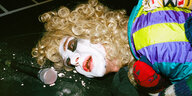 Ein weiß geschminkter Clown mit blonder Locken-Perrücke und Goldzähnen liegt neben einem Mirko mit dem Kopf auf dem Boden