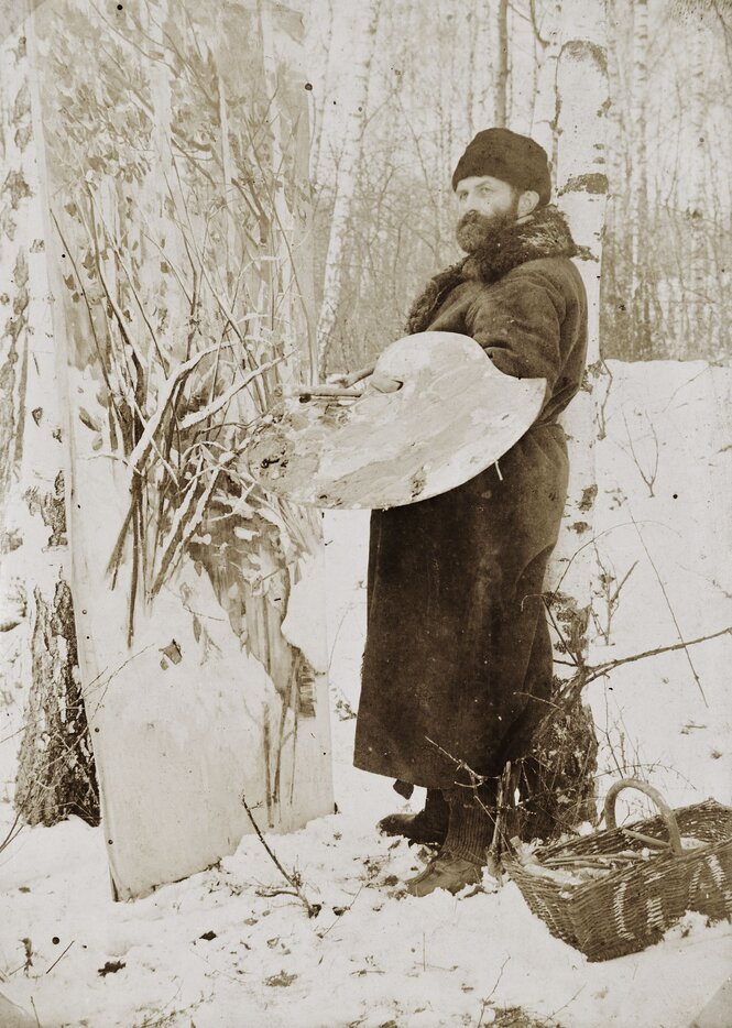 Eine alte schwarzweiße Fotografie, es liegt Schnee, Porträt eines Mannes, der mit Wintermantel und Mütze, Pinsel und Palette vor einer Leinwand steht, die an einer Birke lehnt.