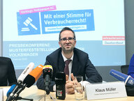 vzbv-Chef Klaus Müller sitzt vor einer Wand mit dem Logo des Bundesverbands der Verbraucherzentralen
