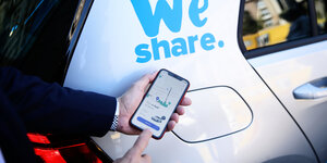 Ein Smartphone mit einer Carsharing App wird vor ein Fahrzeug gehalten.