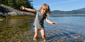 Ein Kind steht mit dem Füßen an einer felsigen Küste im Wasser.
