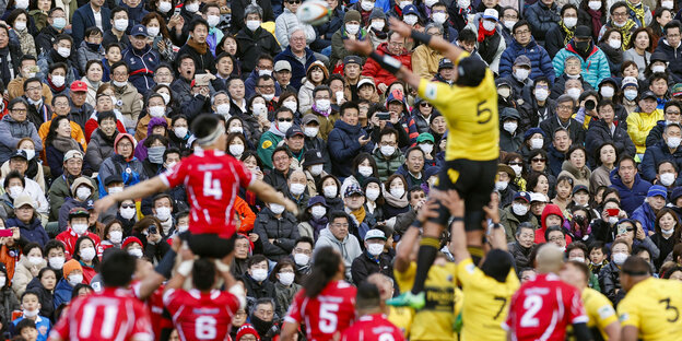 eine voll besetzte Tribühne bei einem Rugbyspiel, viele Zuschauer tragen einen Mundschutz